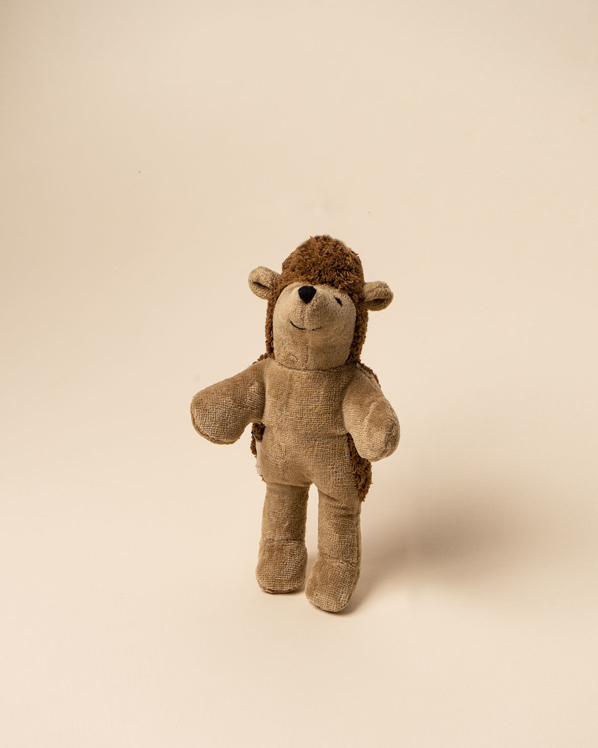 A well-loved brown and tan Senger Naturwelt stuffed hedgehog, handmade in Germany, stands against a plain, light beige background. The toy shows signs of wear, suggesting it is cherished.